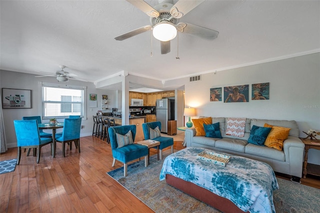 living area featuring baseboards, visible vents, light wood-style flooring, ceiling fan, and crown molding