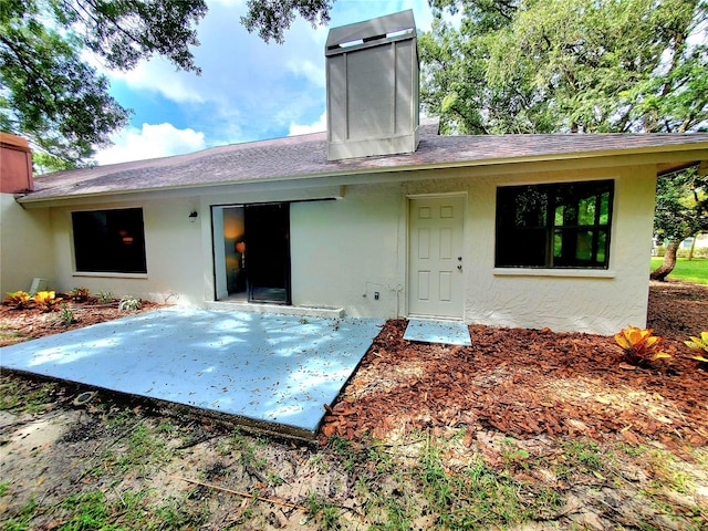 rear view of house with a shingled roof, a patio, a chimney, and stucco siding