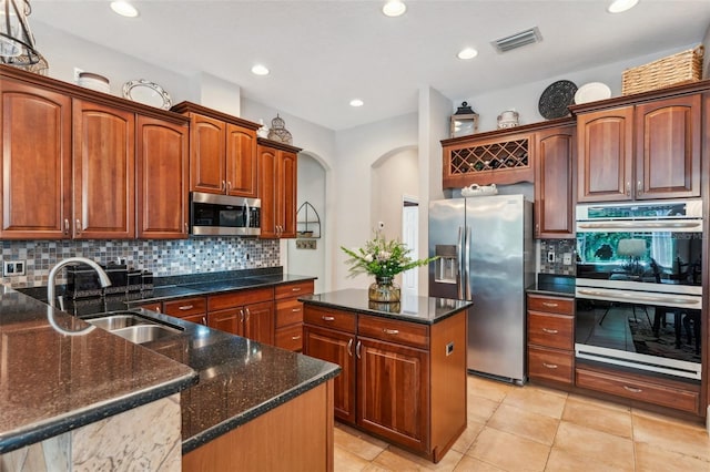 kitchen with sink, a center island with sink, dark stone counters, stainless steel appliances, and backsplash