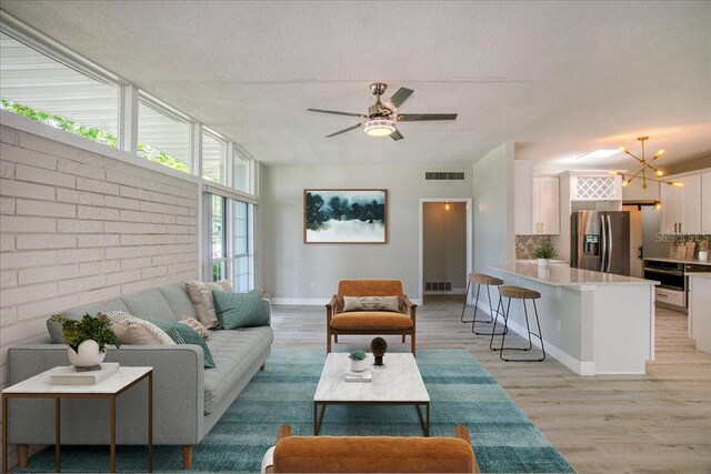 living room featuring ceiling fan with notable chandelier, brick wall, light wood-type flooring, and a textured ceiling