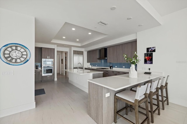 kitchen with tasteful backsplash, a breakfast bar area, appliances with stainless steel finishes, a tray ceiling, and wall chimney range hood