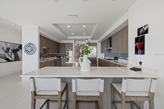 kitchen featuring tasteful backsplash, a kitchen bar, a raised ceiling, wall chimney exhaust hood, and stainless steel appliances