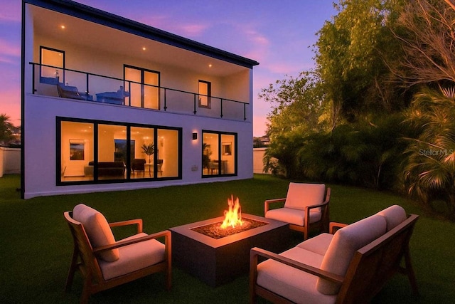 back of property at dusk with an outdoor living space with a fire pit, a balcony, a yard, and stucco siding