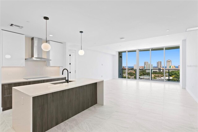 kitchen featuring a center island with sink, wall chimney range hood, dark brown cabinetry, decorative light fixtures, and white cabinetry