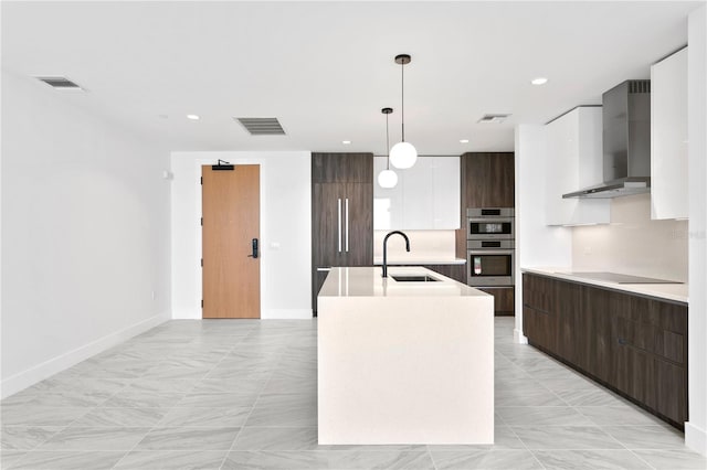 kitchen featuring white cabinetry, sink, wall chimney exhaust hood, hanging light fixtures, and an island with sink