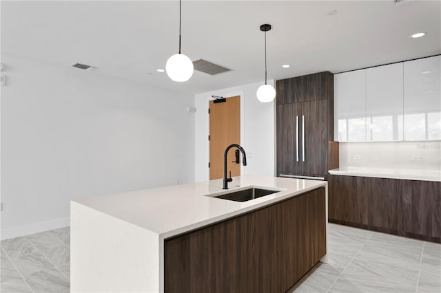 kitchen with paneled fridge, sink, pendant lighting, white cabinetry, and an island with sink