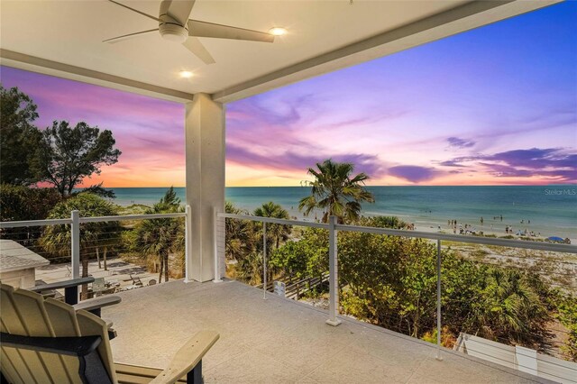 balcony at dusk with a water view, ceiling fan, and a beach view