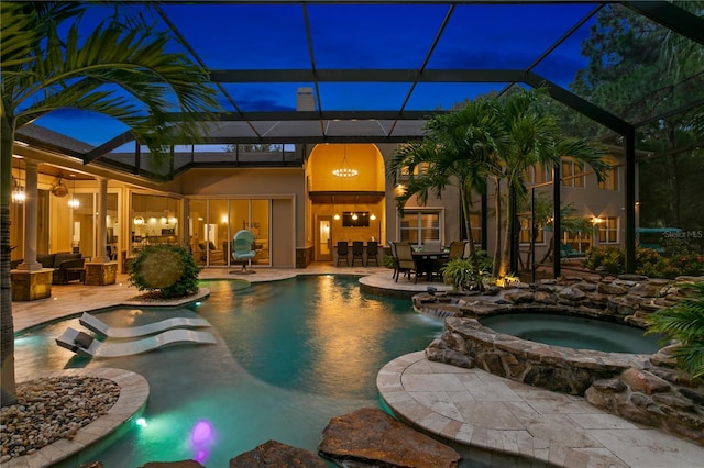 pool at night featuring glass enclosure, a patio area, and an in ground hot tub