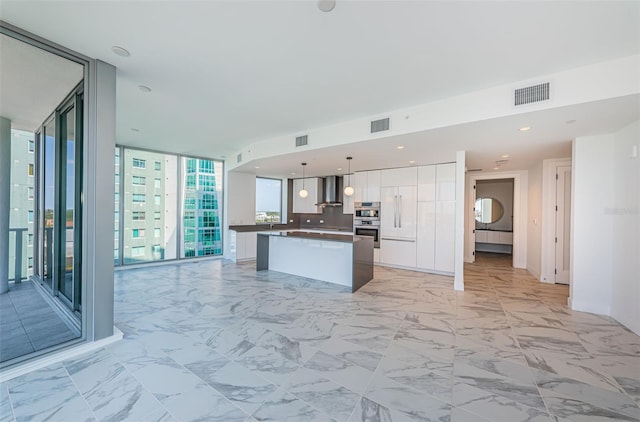 kitchen featuring backsplash, a kitchen island, wall chimney range hood, white cabinetry, and hanging light fixtures