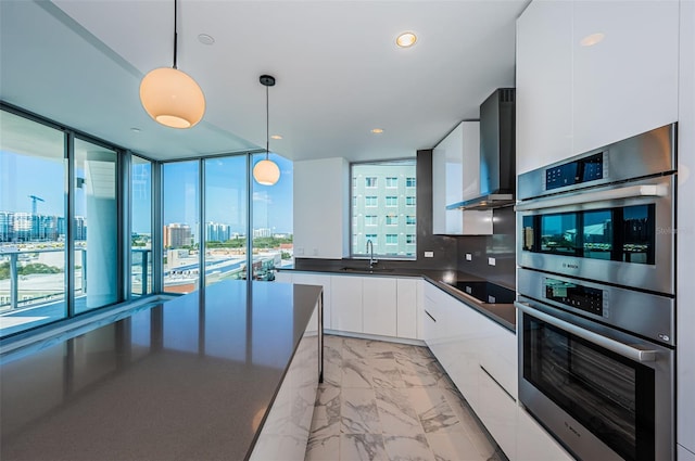 kitchen with backsplash, white cabinets, wall chimney range hood, hanging light fixtures, and stainless steel double oven