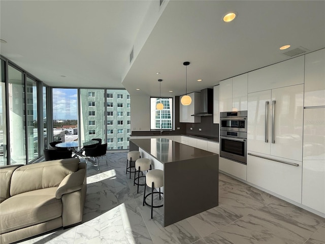 kitchen featuring wall chimney exhaust hood, stainless steel double oven, a center island, white cabinetry, and hanging light fixtures