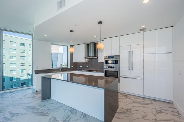 kitchen with dark countertops, wall chimney exhaust hood, marble finish floor, white cabinetry, and pendant lighting