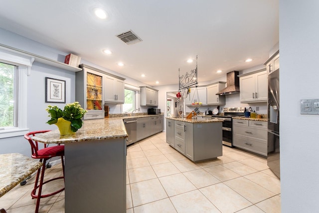 kitchen featuring gray cabinets, an island with sink, wall chimney range hood, appliances with stainless steel finishes, and light tile patterned floors