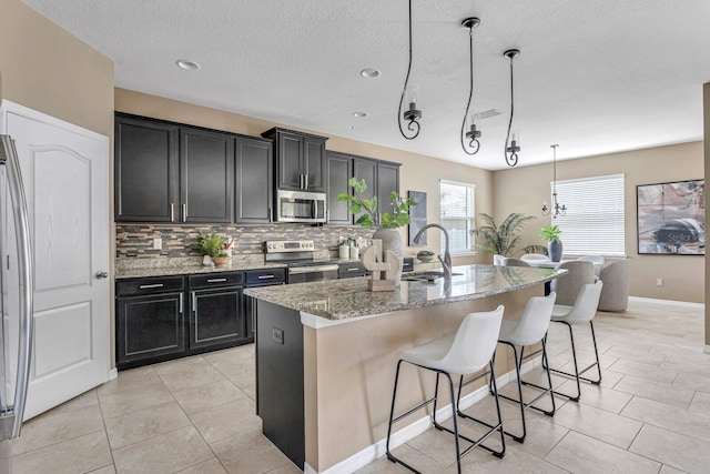 kitchen featuring sink, hanging light fixtures, light stone counters, a center island with sink, and appliances with stainless steel finishes