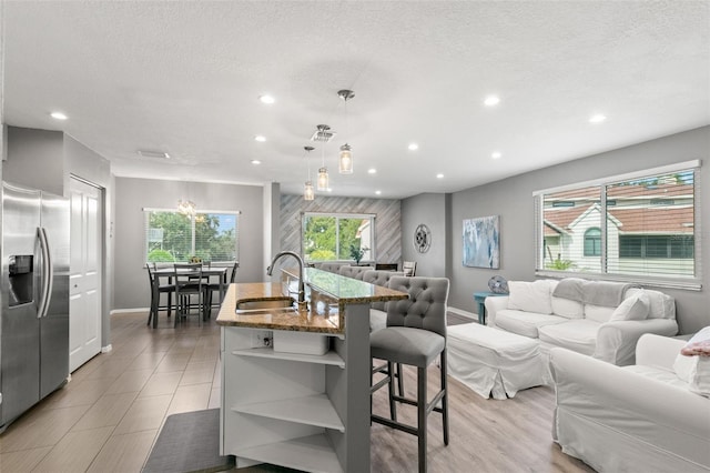 kitchen featuring light stone counters, a kitchen island with sink, a sink, hanging light fixtures, and stainless steel fridge with ice dispenser