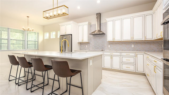 kitchen with appliances with stainless steel finishes, a kitchen island with sink, light tile patterned floors, decorative backsplash, and wall chimney exhaust hood