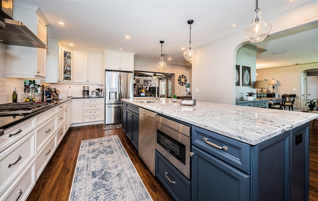 kitchen featuring light stone countertops, wall chimney exhaust hood, blue cabinetry, an island with sink, and stainless steel appliances