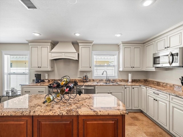 kitchen with a kitchen island, custom range hood, appliances with stainless steel finishes, sink, and light stone counters