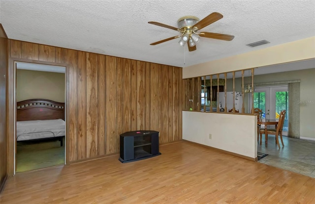 living room with a textured ceiling, ceiling fan, wooden walls, french doors, and light hardwood / wood-style floors