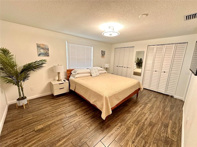 bedroom with a textured ceiling, visible vents, baseboards, dark wood finished floors, and two closets