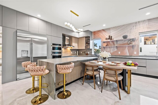 kitchen with decorative light fixtures, a breakfast bar area, wall chimney exhaust hood, appliances with stainless steel finishes, and gray cabinetry