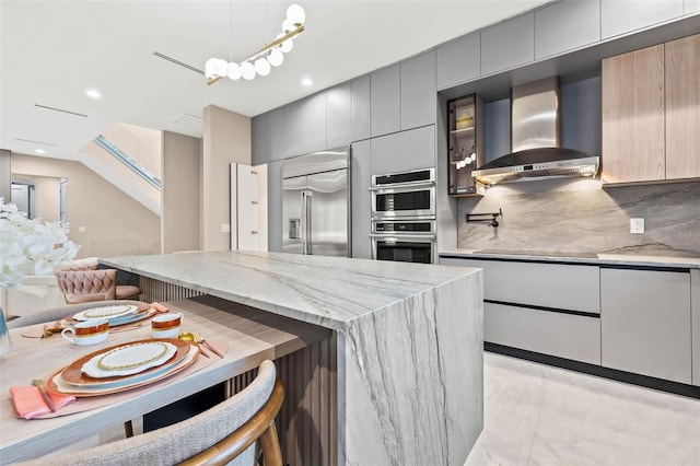 kitchen with gray cabinetry, backsplash, appliances with stainless steel finishes, light stone countertops, and wall chimney exhaust hood