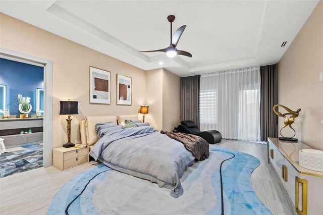 bedroom with light wood-type flooring, a raised ceiling, ceiling fan, and ensuite bath