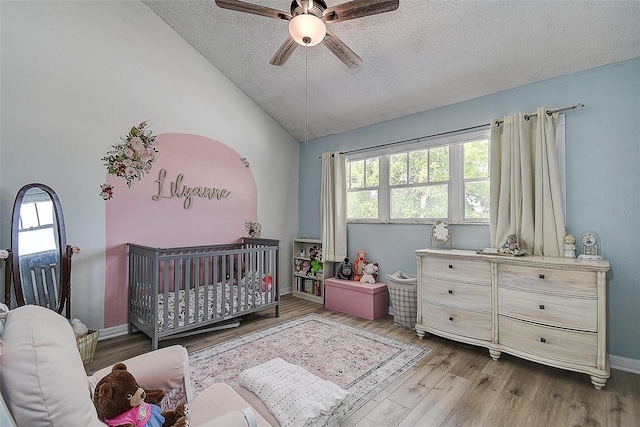 bedroom with ceiling fan, light wood-type flooring, a crib, vaulted ceiling, and a textured ceiling