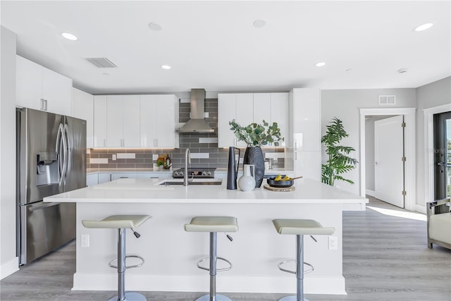 kitchen featuring a breakfast bar, visible vents, stainless steel fridge with ice dispenser, wall chimney exhaust hood, and a center island with sink