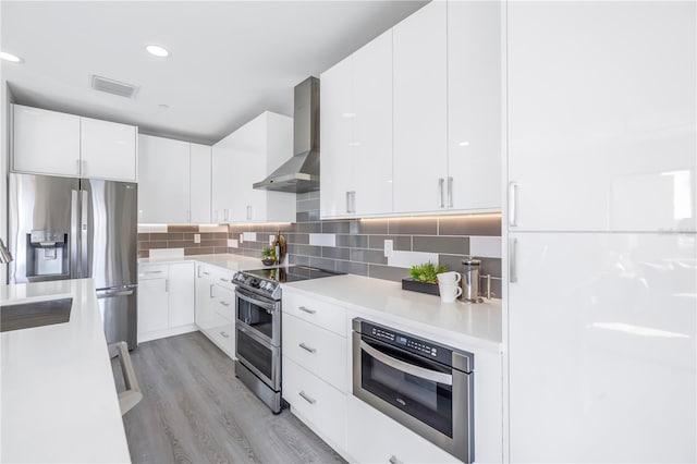 kitchen featuring wall chimney exhaust hood, stainless steel appliances, light countertops, white cabinetry, and backsplash