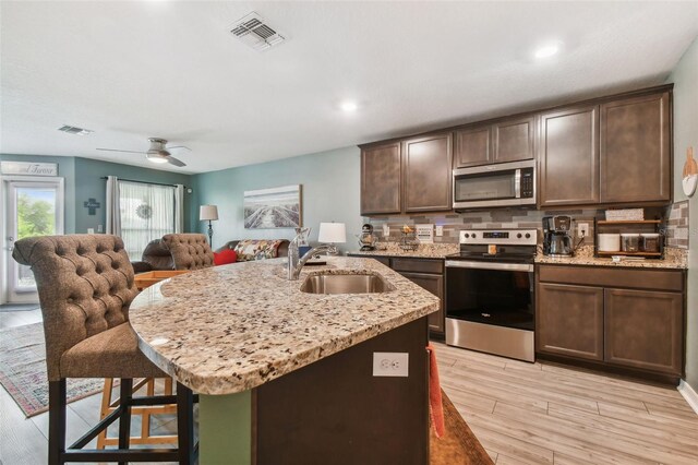 kitchen with appliances with stainless steel finishes, decorative backsplash, dark brown cabinets, an island with sink, and ceiling fan