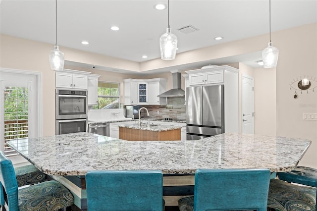 kitchen featuring a large island with sink, pendant lighting, tasteful backsplash, stainless steel appliances, and wall chimney exhaust hood