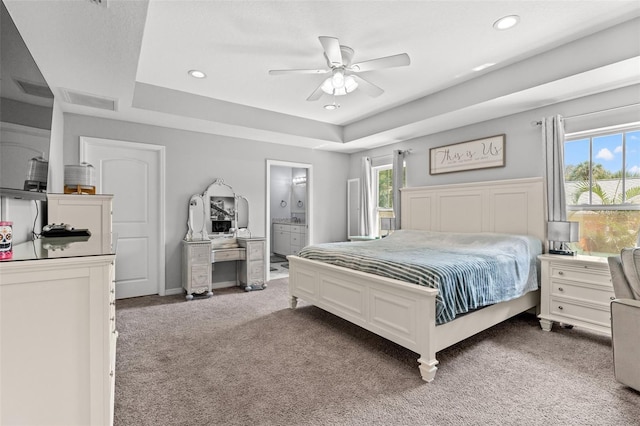 carpeted bedroom featuring a raised ceiling, ensuite bath, ceiling fan, and multiple windows