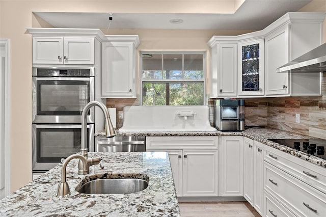 kitchen featuring wall chimney range hood, stainless steel double oven, decorative backsplash, and white cabinetry