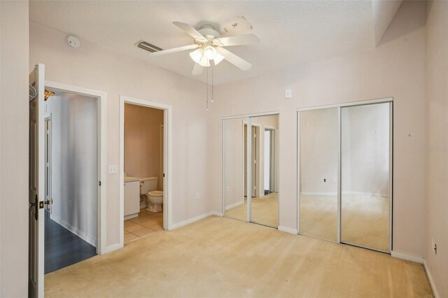 unfurnished bedroom with light colored carpet, two closets, ensuite bath, and ceiling fan