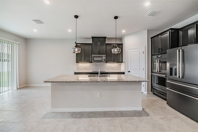 kitchen with appliances with stainless steel finishes, light stone countertops, an island with sink, light tile patterned flooring, and decorative backsplash