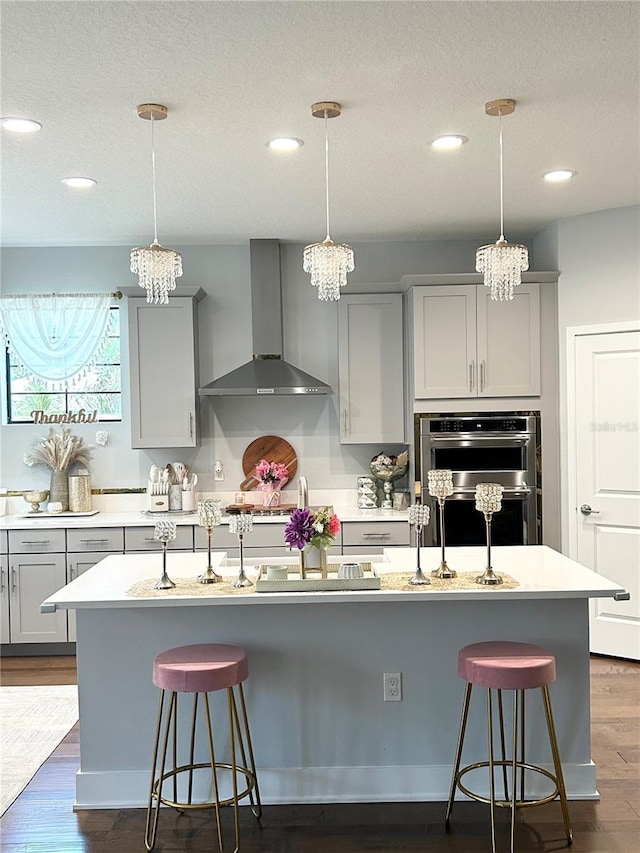 kitchen with light countertops, gray cabinetry, stainless steel double oven, wall chimney range hood, and a kitchen bar