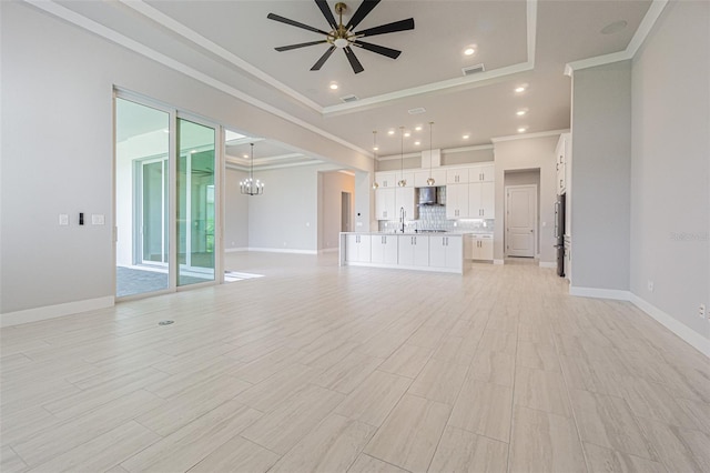 unfurnished living room with ceiling fan with notable chandelier, a raised ceiling, sink, ornamental molding, and light wood-type flooring