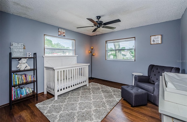 bedroom with a nursery area, ceiling fan, dark wood-type flooring, and a textured ceiling