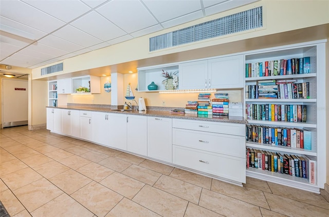 kitchen with light stone countertops, light tile patterned floors, sink, white cabinetry, and a drop ceiling