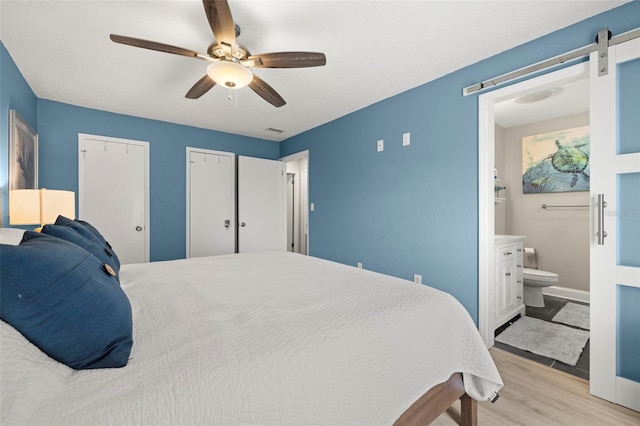 bedroom with multiple closets, light wood-type flooring, a barn door, ceiling fan, and connected bathroom