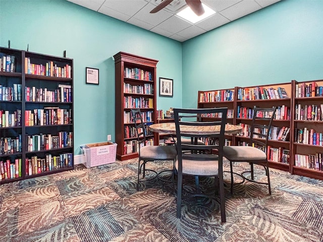 living area featuring a paneled ceiling, wall of books, ceiling fan, and baseboards