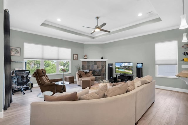 living area with baseboards, light wood-style floors, a tiled fireplace, a raised ceiling, and crown molding