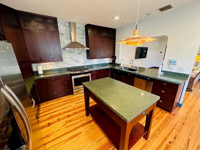 kitchen with appliances with stainless steel finishes, light wood-type flooring, decorative light fixtures, sink, and wall chimney range hood