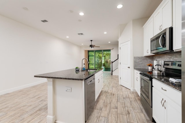 kitchen featuring a kitchen island with sink, light wood-type flooring, dark stone countertops, stainless steel appliances, and ceiling fan