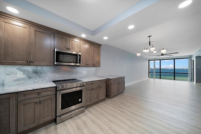 kitchen with ceiling fan with notable chandelier, backsplash, stainless steel appliances, light wood-type flooring, and a water view