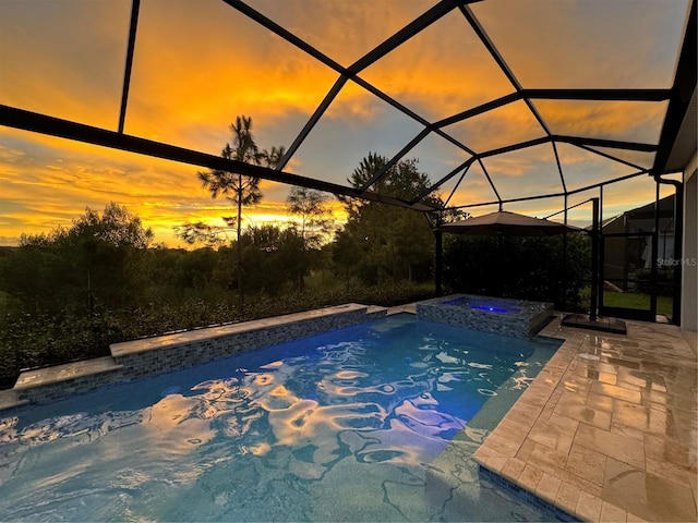 pool at dusk featuring glass enclosure, an in ground hot tub, and a patio