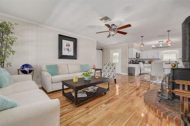 living room with light wood-type flooring, crown molding, a textured ceiling, a wood stove, and ceiling fan