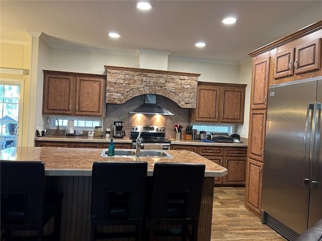 kitchen featuring backsplash, appliances with stainless steel finishes, light hardwood / wood-style floors, an island with sink, and wall chimney range hood