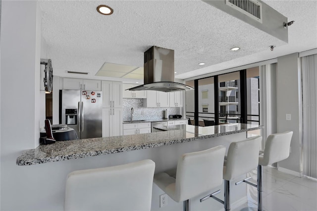 kitchen featuring light tile floors, stainless steel fridge with ice dispenser, white cabinets, backsplash, and island range hood